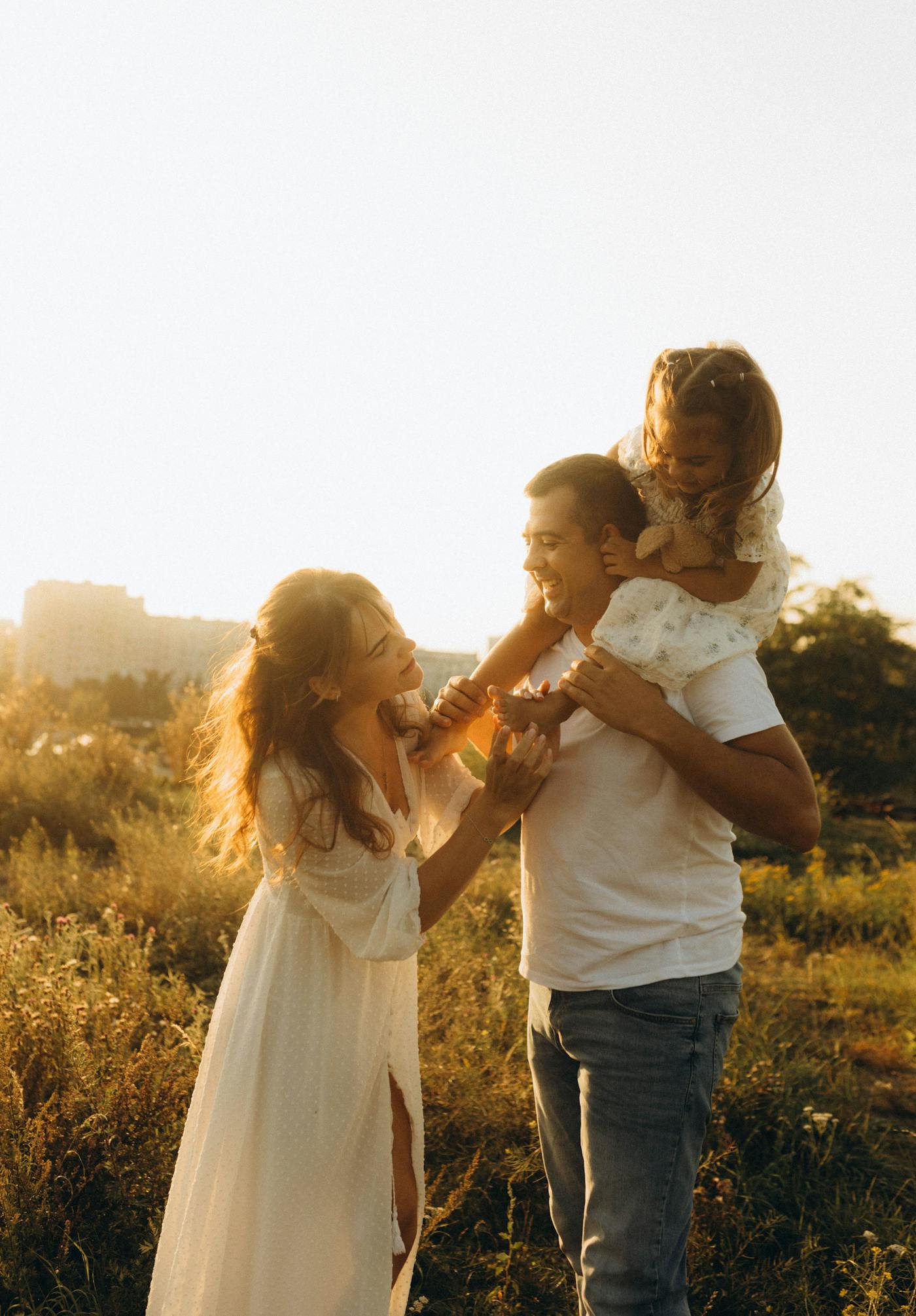 Parents laughing with their daughter, who rides her dad’s shoulders in a sunlit field