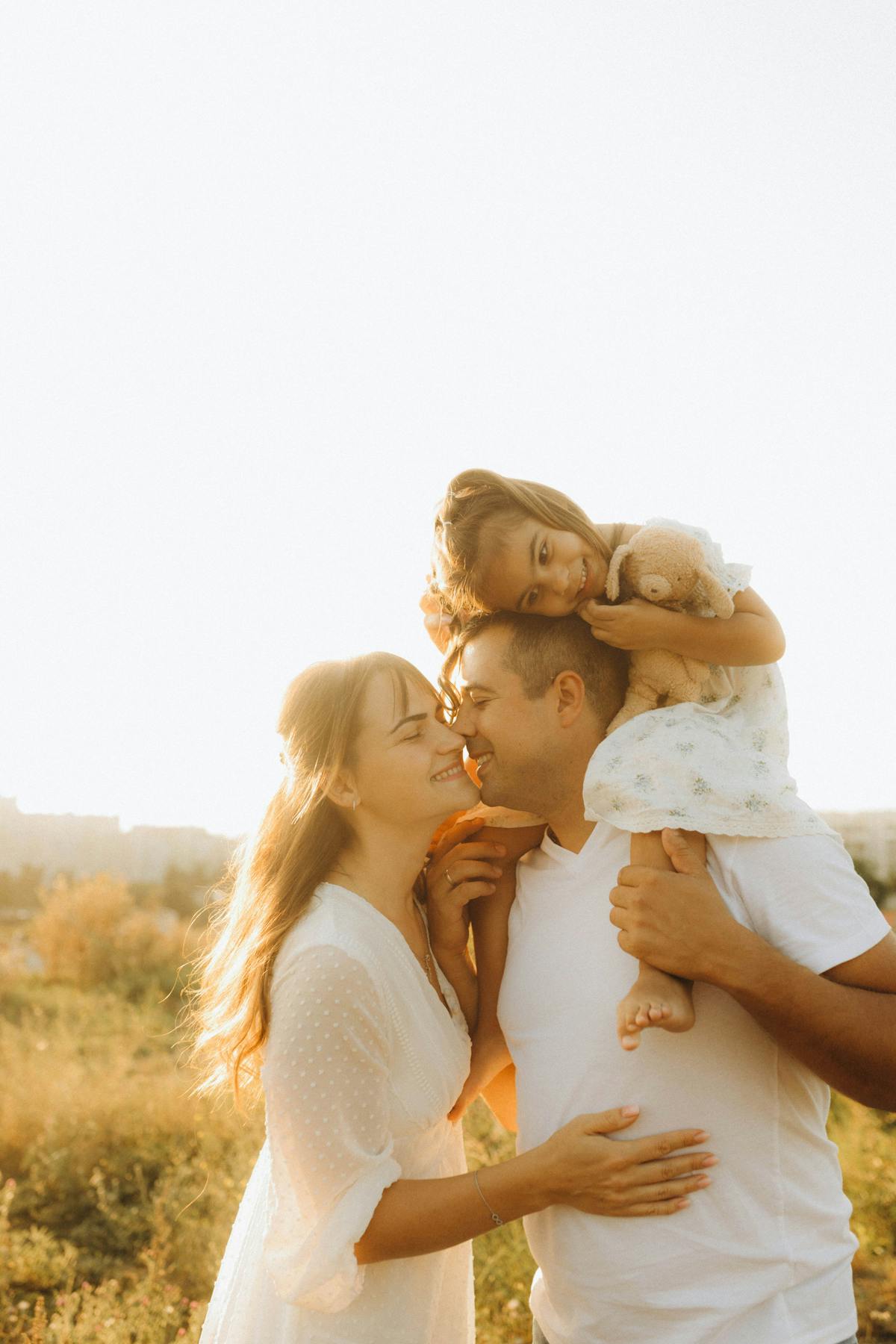 Girl with a teddy bear on her dad’s shoulders while her parents touch noses